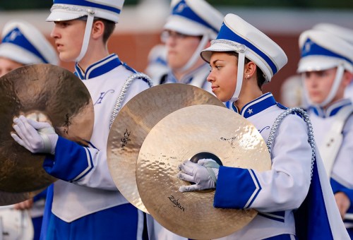A young female cymbal player in a blue and white marching band uniform stands among fellow musicians, focused and ready.