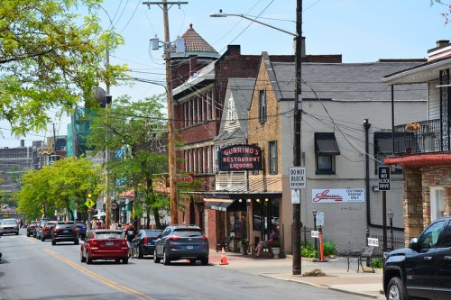 A street view featuring buildings, restaurants, and parked cars under a clear blue sky. Green trees line the road.