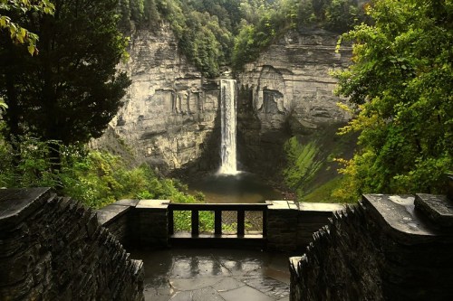 A scenic view of a waterfall cascading into a lush green gorge, framed by trees and stone structures.