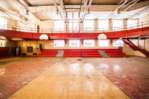 Abandoned gymnasium with red bleachers, empty basketball hoops, and scattered debris on the wooden floor.