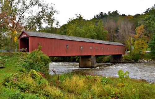 A red covered bridge spans a river, surrounded by trees and autumn foliage.