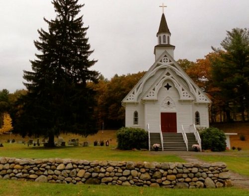 A white church with a steeple, surrounded by trees and a stone wall, set against a cloudy sky.
