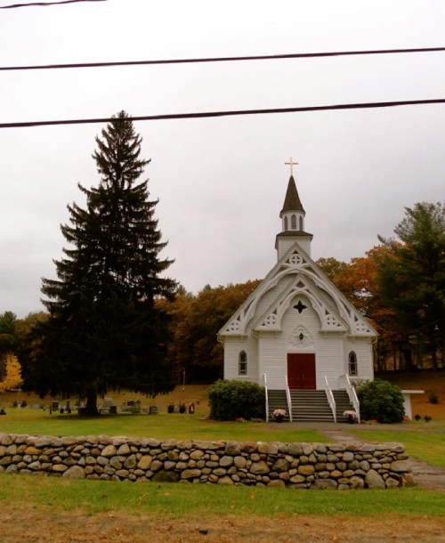 A small white church with a steeple, surrounded by trees and a stone wall, set against a cloudy sky.