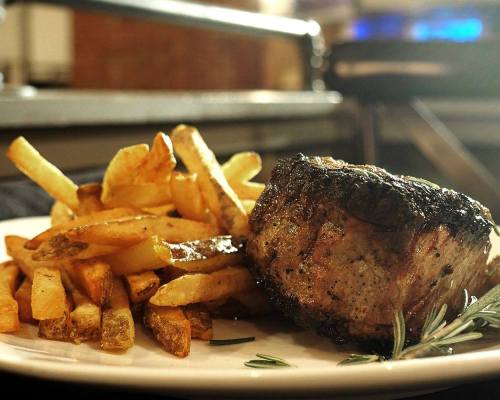 A plate of grilled steak beside a serving of golden French fries, garnished with a sprig of rosemary.