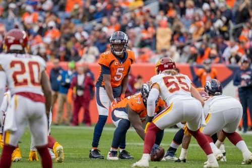 A quarterback in an orange jersey prepares to pass during an NFL game, with players from both teams in action.
