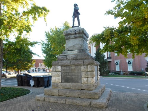 A stone monument with a soldier statue on top, surrounded by trees and buildings in a town square.