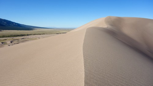A vast sandy dune under a clear blue sky, with rolling hills and distant mountains in the background.