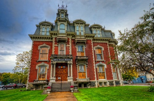 Historic red brick mansion with ornate architecture, featuring a grand entrance and boarded windows, set in a green landscape.
