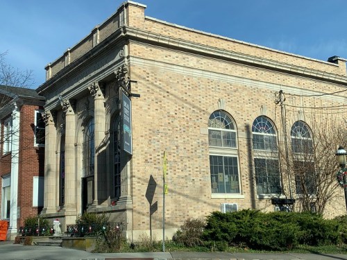 Historic brick building with large windows and columns, surrounded by greenery and a clear blue sky.