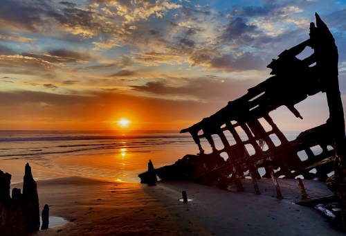 A shipwreck silhouette against a vibrant sunset over the ocean, with clouds reflecting warm colors in the sky.
