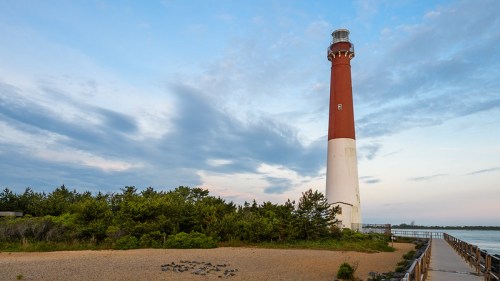 A red and white lighthouse stands near a sandy beach, surrounded by greenery under a cloudy sky.