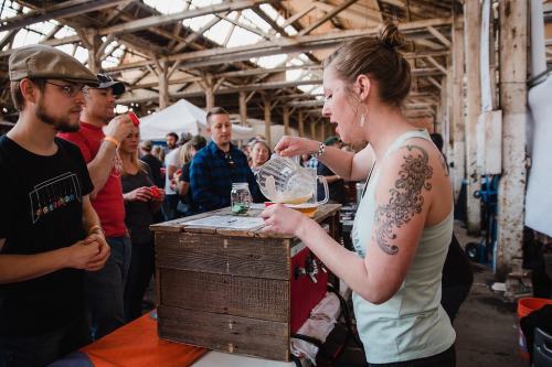 A woman pours a drink from a pitcher at a market, while onlookers watch in a rustic indoor setting.