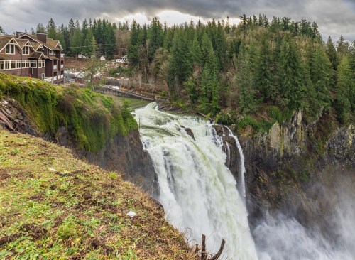 A powerful waterfall cascades down rocky cliffs, surrounded by lush green trees and a rustic building nearby.