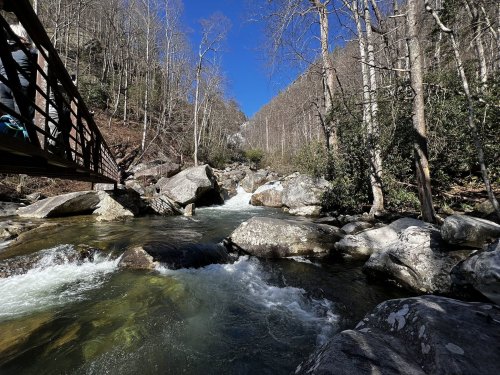 A serene river flows through rocky terrain, surrounded by trees under a clear blue sky. A bridge is visible in the background.