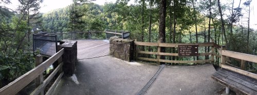 A scenic overlook surrounded by trees, featuring a wooden railing and benches, with a view of distant hills.