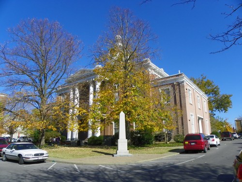 A brick building with columns, surrounded by trees with autumn leaves, and parked cars in a sunny blue sky.