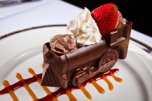 A chocolate dessert shaped like a train, topped with whipped cream and a strawberry, on a plate with sauce drizzles.