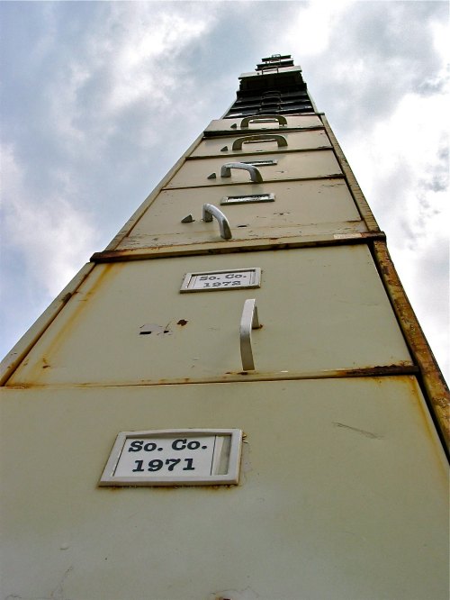 A tall, rusted structure with metal rungs and plaques labeled "So. Co. 1971" against a cloudy sky.