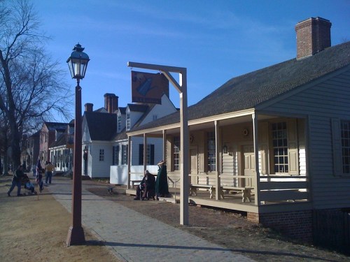 Historic building with a porch, lamp post, and sign, set against a clear blue sky. People walk along the path.
