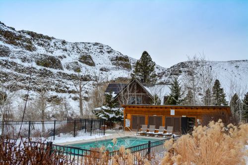 A cozy cabin near a snowy landscape, featuring a pool surrounded by a fence and winter vegetation.