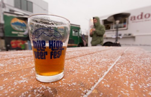 A glass of beer sits on a wooden table, with a food truck in the background and light snow falling.