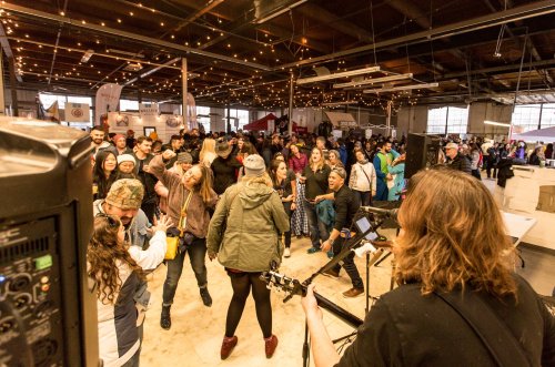 A lively crowd dances at an indoor event, with a musician playing guitar in the foreground and festive lights overhead.