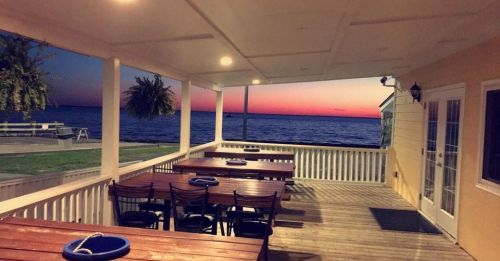 A waterfront patio with wooden tables and chairs, overlooking a colorful sunset over the water.