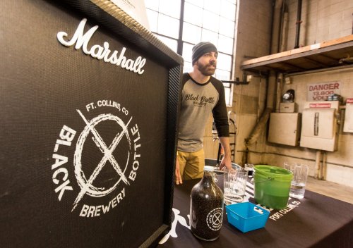 A man stands behind a table with brewery branding, featuring bottles and glasses in a warehouse setting.