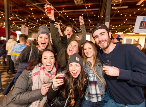 A group of happy friends smiling and holding drinks at a lively indoor event with festive lighting.