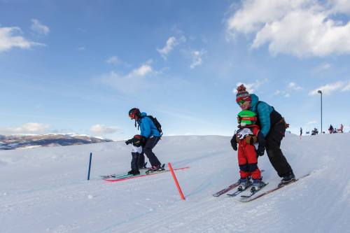 A parent helps a child ski on a snowy slope, with another skier in the background under a blue sky.