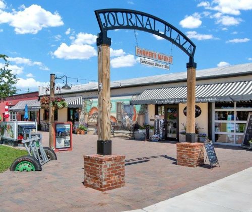 A vibrant outdoor plaza with shops, colorful murals, and a sign for "Journal Plaza" under a blue sky.