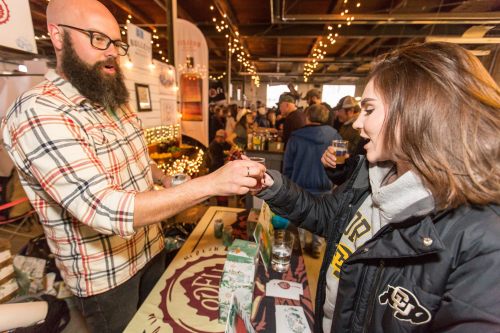 A bearded man hands a drink to a woman at a bustling event with festive lighting and booths in the background.