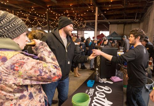 A lively indoor event with people enjoying drinks at a bar, string lights overhead, and a vendor serving beer.