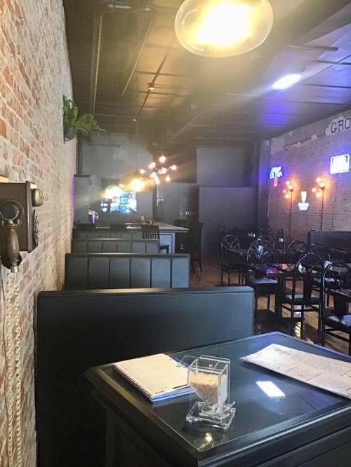 Interior of a restaurant featuring brick walls, black booths, and pendant lighting, with a menu on a table.
