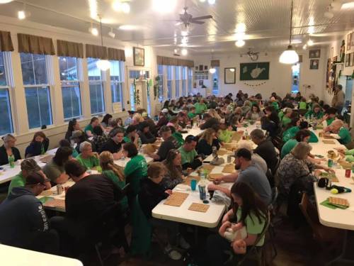 A large group of people in green shirts gathered around tables, playing bingo in a well-lit community hall.
