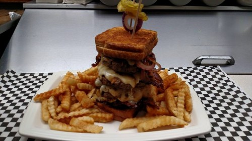 A towering burger with bacon, cheese, and grilled onions, served with crinkle-cut fries on a checkered plate.