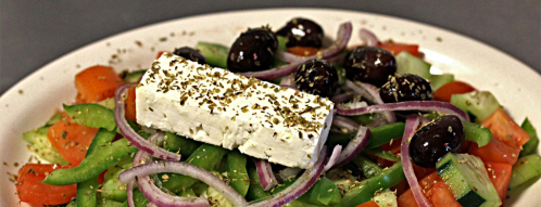 A colorful salad with tomatoes, cucumbers, green peppers, red onions, olives, and a block of feta cheese on top.