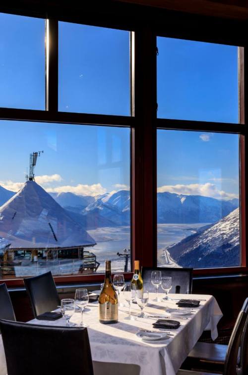 A dining table set with wine glasses and a bottle, overlooking a snowy mountain landscape through large windows.
