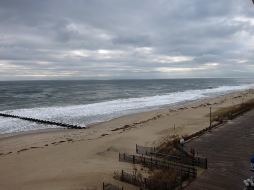 A cloudy beach scene with gentle waves, sandy shore, and wooden posts extending into the water.