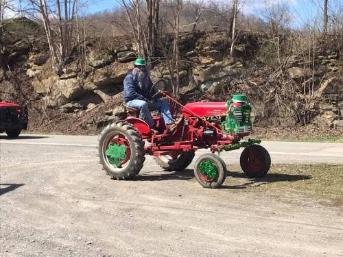 A person with a beard rides a red tractor decorated for St. Patrick's Day, surrounded by trees and gravel.