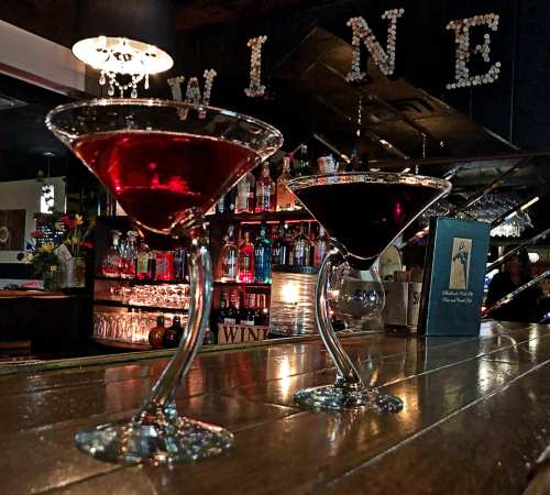 Two elegant cocktails in martini glasses, one red and one dark, on a wooden bar with a "WINE" sign in the background.