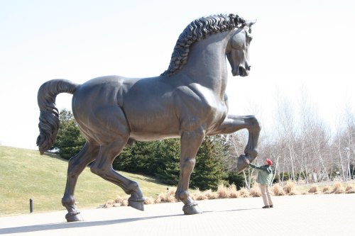 A person stands next to a large bronze horse statue, showcasing its impressive size against a clear sky.
