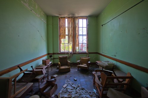 Abandoned room with green walls, scattered furniture, and sunlight filtering through dusty windows.