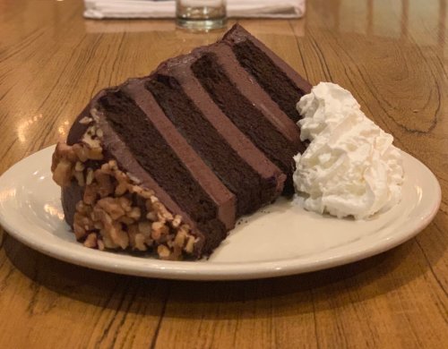A slice of chocolate cake with layers and a side of whipped cream on a white plate, resting on a wooden table.