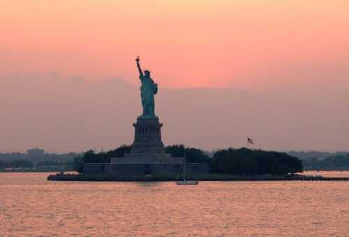 The Statue of Liberty stands tall against a sunset, with a sailboat in the foreground and a calm water surface.