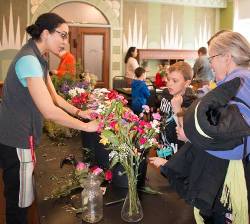 A woman demonstrates flower arranging to children and an adult at a community event, surrounded by colorful flowers.