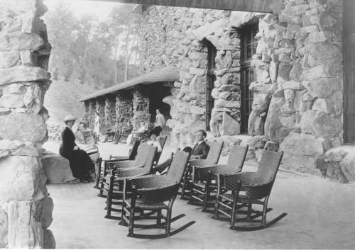 A historic black-and-white photo of people sitting in rocking chairs on a porch made of stone.