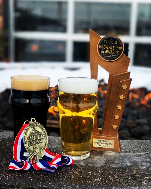 Two glasses of beer, one dark and one light, alongside a trophy and a medal, set against a fire pit background.