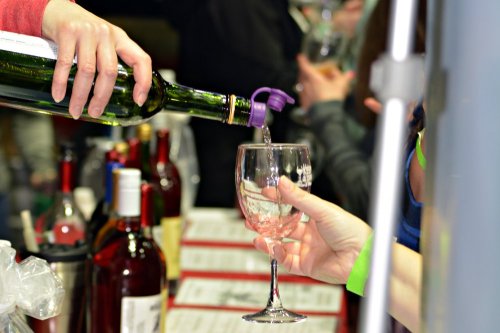 A hand pours wine from a bottle into a glass at a busy bar or event, with bottles and people in the background.