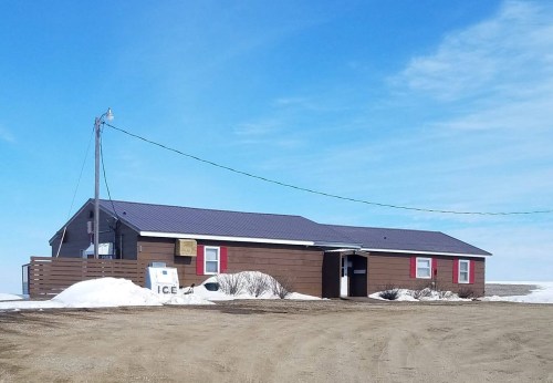 A brown house with a blue roof, red shutters, and snow around it, set against a clear blue sky.